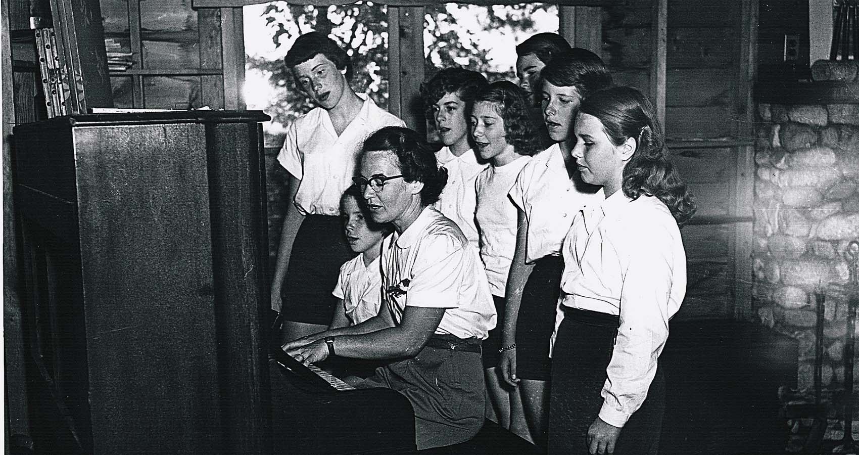 Historical photo of campers singing with director around a piano