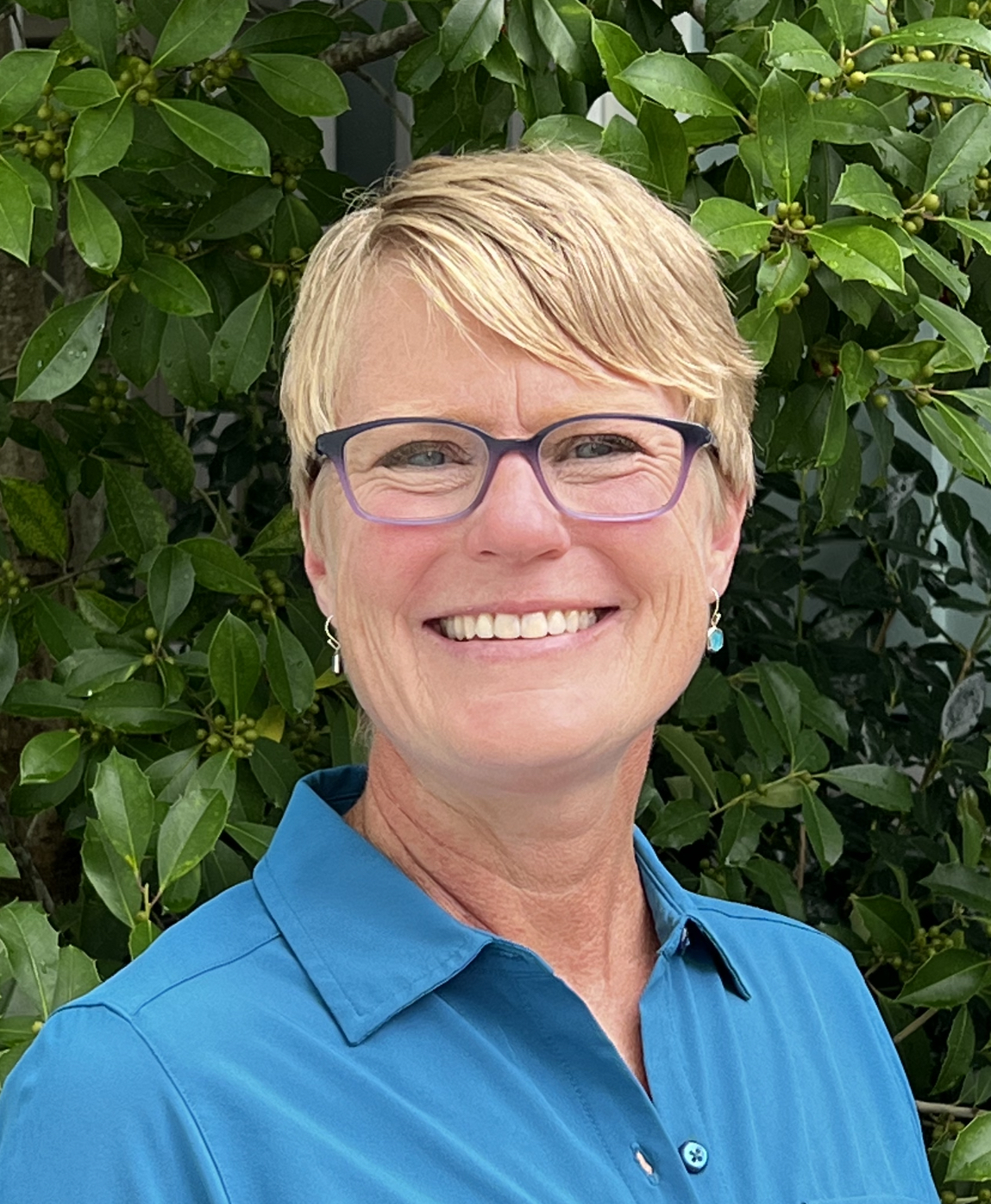 Photo of Kim Aycock smiling wearing thick frames positioned in front of a green bush