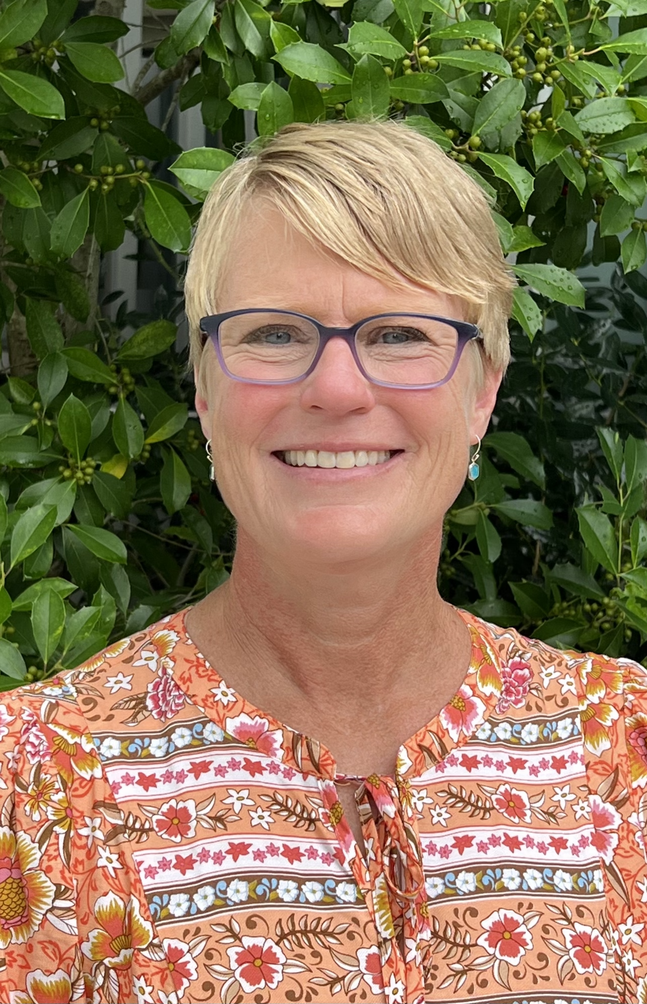 Photo of Kim Aycock smiling wearing thick frames positioned in front of green bushed