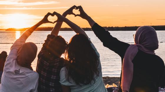 Four people facing water on beach holding hands making heart shapes