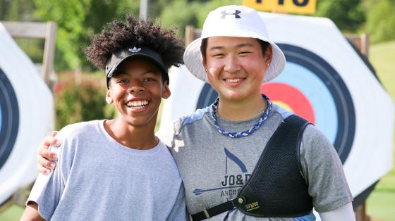 Two smiling young people posing together at an archery range, with colorful circular targets visible in the background