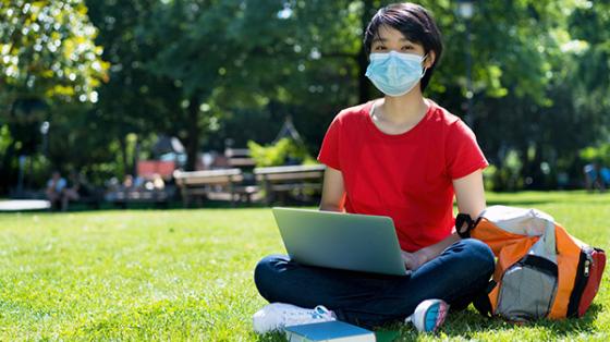 college student wearing mask sitting in grass with laptop