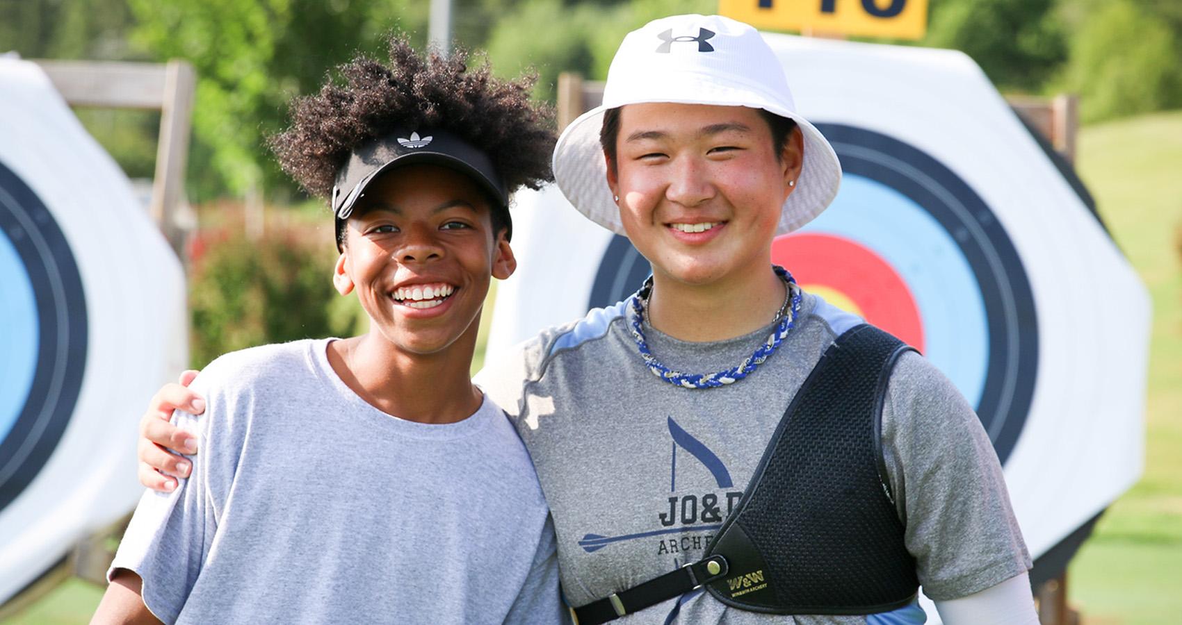 Two smiling young people posing together at an archery range, with colorful circular targets visible in the background