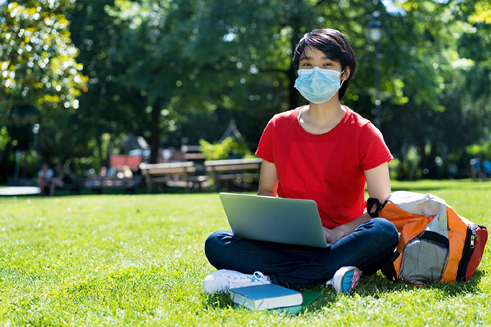 college student wearing mask sitting in grass with laptop