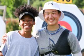 Two smiling young people posing together at an archery range, with colorful circular targets visible in the background